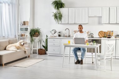 Engelli bir Afrikalı Amerikalı adam bir masada oturuyor, yanında sadık Labrador rehber köpeği ile dizüstü bilgisayarında yazıyor..