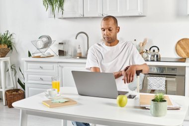A diverse African American man with myasthenia gravis sits at a kitchen table, engrossed in his laptop. clipart