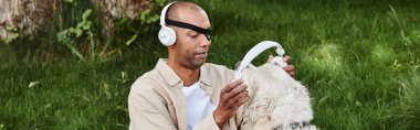 Miyastenia gravis sendromu olan Afro-Amerikalı bir adam, kulaklık takan bir Labrador köpeğiyle çimenlerde dinleniyor..