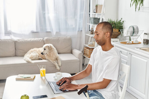 A disabled African American man sits at a table using a laptop, accompanied by his Labrador dog.