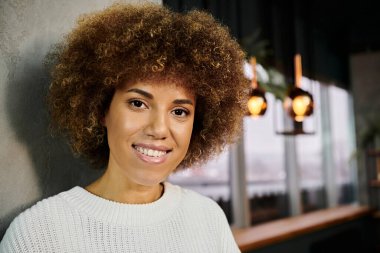 Gülümseyen Afrikalı Afro saçlı bir kadın modern bir kafede kameraya poz veriyor, mutluluk saçıyor..