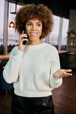 Beyaz kazak giyen Afro-Amerikalı bir kadın modern bir kafe ortamında cep telefonuyla sohbet ediyor..