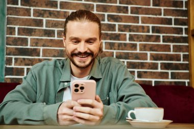 Mutlu adam modern bir kafede bir masada oturuyor ve cep telefonunu kullanmaya odaklanıyor..