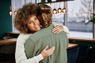 An African American woman tenderly hugs a man in a modern restaurant setting. clipart