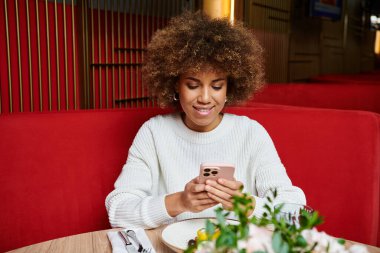 A stylish African American woman immerses in her phone while seated at a cafe table. clipart