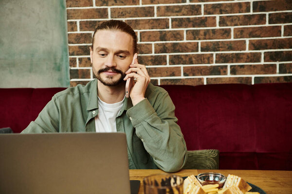 Мужчина сидит за столом в современном кафе, разговаривает по мобильному телефону.