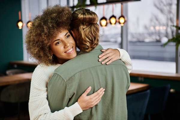 An African American woman tenderly hugs a man in a modern restaurant setting.