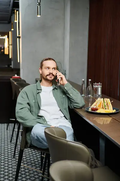 Mężczyzna Siedzący Przy Stole Pochłonięty Telefonem Otoczony Nowoczesną Atmosferą Kawiarni — Zdjęcie stockowe