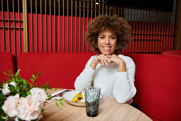 stock image An African American woman sits at a modern cafe table, gracefully enjoying a plate of delicious food.