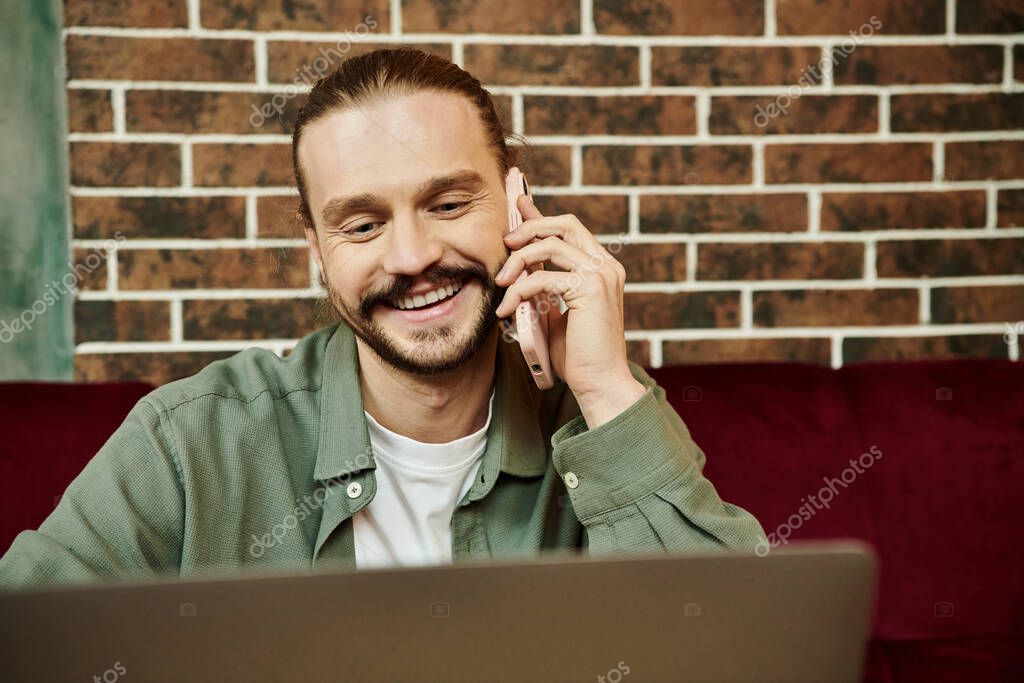 A man sitting in front of a laptop, engaged in a conversation on his cell phone.