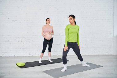 İki hamile kadın yoga minderlerinin üzerinde güvenle duruyor, ebeveynlik kurslarında koçlarının rehberliğinde alıştırma yapıyorlar..