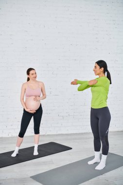 Hamile bir kadın yoga minderinde dururken diğer hamile kadın arka planda ebeveyn kursunda birlikte pratik yapıyor..