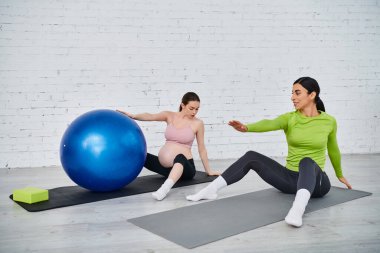 İki kadın, biri hamile, veli kursu sırasında yoga minderlerinde oturuyorlar..