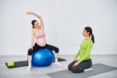 Hamile bir kadın antrenörüyle veli kurslarında mavi bir fitness topunun tepesinde otururken denge ve güç bulur..