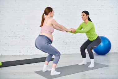 İki kadın, biri hamile, yoga minderinin üzerinde zarif bir şekilde duruyor, dingin bir ortamda güç ve denge yayıyor.