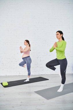 İki kadın, biri hamile, yoga pozisyonlarında zarifçe poz veriyor, beyaz tuğla bir duvarın önünde huzur ve güç buluyor..