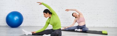 Hamile bir kadın, ebeveyn kursları sırasında koçuyla yoga yapıyor. Her ikisi de yoga minderleri üzerinde rahat bir ortamda oturuyor..