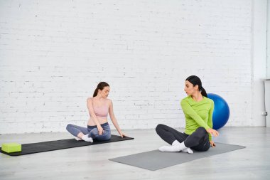 Hamile bir kadın, ebeveyn kursu sırasında koçuyla egzersiz yapıyor. Yoga minderleri üzerinde oturuyor..
