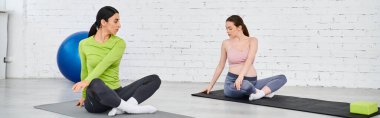 İki kadın, biri hamile, ebeveyn kursu sırasında yoga minderlerinin üzerinde rahatça otururlar..