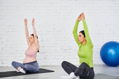Tuğla duvarın önünde yoga yapan iki kadın; biri hamile, ebeveyn kursu sırasında koçu tarafından rehberlik ediliyor..