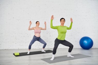 Koçunun rehberliğinde hamile bir kadın huzurlu bir ortamda minderlerin üzerinde yoga yapıyor..
