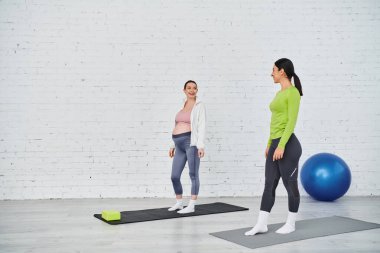 İki kadın, biri hamile, spor salonunda yoga minderlerinin üzerinde duruyor, ebeveyn kursu sırasında sakin bir farkındalık ve hareket anı yaşıyor..