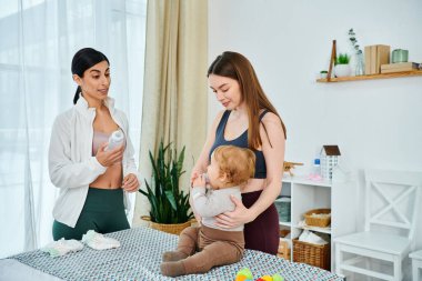 Genç bir anne bebeğini kollarına alır. Bakıcı da yanında bir şişe taşır. Destek ve rehberlik sağlar..