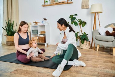 Genç bir anne, koçu tarafından bir ebeveynlik kursunda rehberlik edilen bebeğiyle yoga minderinde oturur ve bir anlık huzur ve bağ kurar..