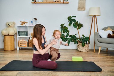 Yoga minderinde oturan genç ve güzel bir anne ebeveynlik kursunda koçunun rehberliğinde bebeğini kucağına alıyor..