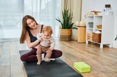 Yoga minderinde oturan genç bir anne, ebeveynlik kursunda koçun rehberliğiyle bebeğini kucaklar..