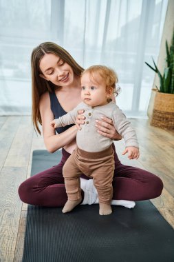 Genç bir anne bebeğini nazikçe yoga minderinde tutarken, ebeveyn kurslarında koçun rehberliğinde evlerinin rahatlığında..