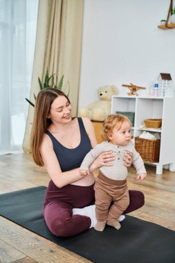 A young, beautiful mother sits on a yoga mat, peacefully cradling her baby with the guidance of her coach at parents courses. clipart