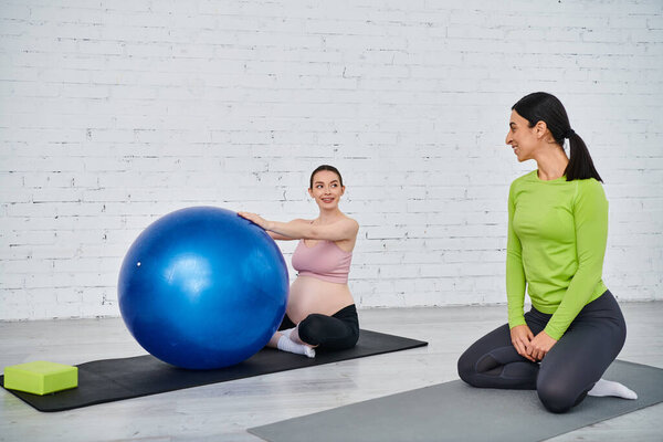 Two women, one pregnant, are seated on yoga mats engaging with a large exercise ball under the guidance of a coach during parents courses.