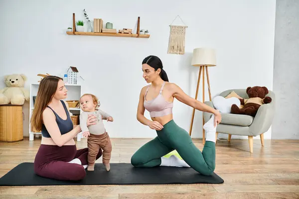 stock image A young beautiful mother is sitting on a yoga mat, gently holding her baby while receiving guidance from her coach.