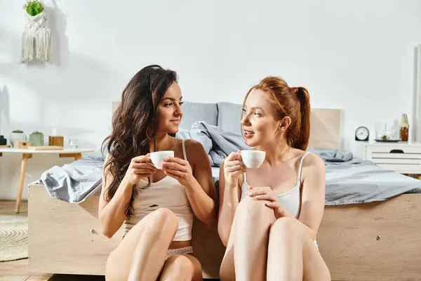 Two elegant women, a loving lesbian couple, sit on a bed enjoying coffee together in a cozy setting.