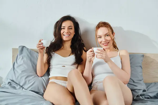 stock image Two women in elegant attire, sitting on a bed, enjoying coffee together.