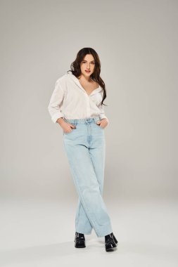 A beautiful plus size woman strikes a pose in a white shirt and blue jeans against a gray backdrop. clipart