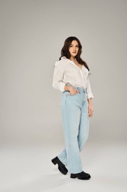 Stylish plus size woman posing confidently in white shirt and blue jeans on a gray backdrop. clipart