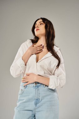 A stunning plus size woman strikes a pose in a trendy white shirt and blue jeans against a neutral gray backdrop. clipart