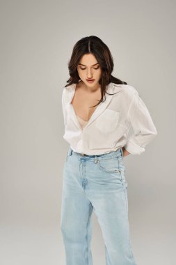 A beautiful plus size woman poses confidently in a stylish white shirt and blue jeans against a gray backdrop. clipart