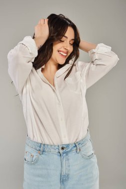 A beautiful plus size woman posing in stylish white shirt and jeans against a gray backdrop. clipart