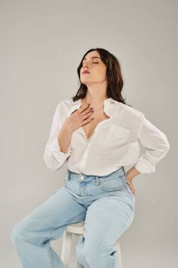 Güzel ve iri yarı bir kadın şık giyinmiş, tabureye oturmuş, ellerini göğsüne koyuyor..