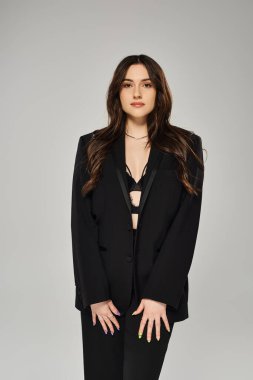 Siyah takım elbiseli, iri yarı, güzel bir kadın gri bir fonda kendine güvenen bir poz veriyor..