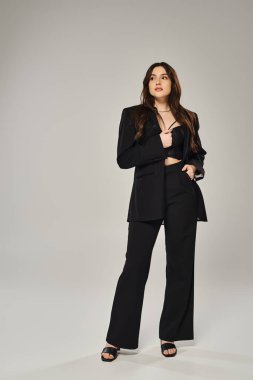 Gri bir zemine karşı siyah bir takım elbiseyle poz verirken kendine güveni ve stili yüksek, güzel bir kadın..