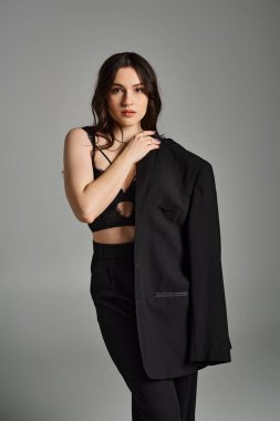 Gri bir arkaplana karşı güvenle poz veren parlak siyah takım elbiseli, iri yarı, güzel bir kadın..