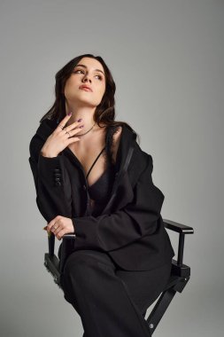 Tarz sahibi bir kadın derin düşüncelere dalmış, çenesi elinde dinleniyor, gri bir arka plana karşı zarif bir şekilde sandalyede oturuyor..
