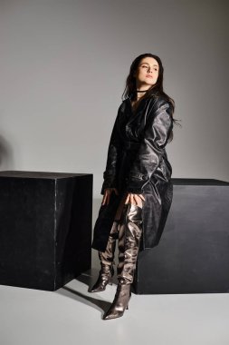 Gri bir zeminde gizemli bir kara kutunun yanında zarif, iri yarı bir kadın duruyor..