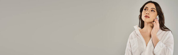 A beautiful plus-size woman in stylish attire gracefully holds her hand to her face against a gray backdrop.