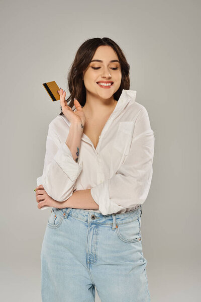 Красивая женщина в стильном наряде, держащая кредитку и улыбающаяся на сером фоне.