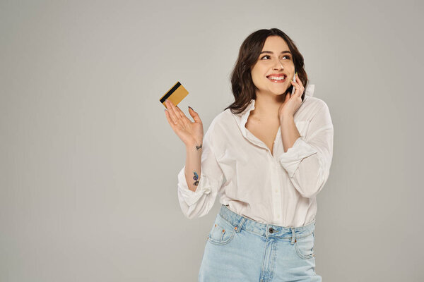 A stylish plus size woman multitasking, holding a credit card and talking on a cell phone against a gray backdrop.